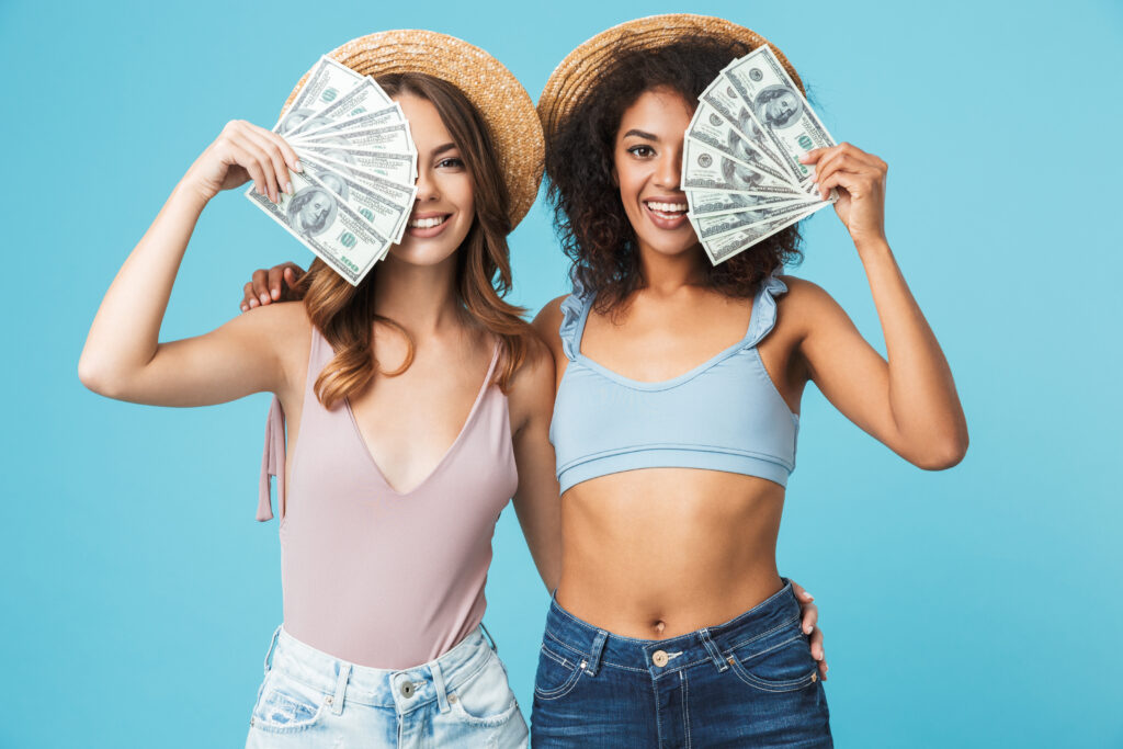 Photo of caucasian and african american women wearing straw hats and summer clothing covering their faces with dollar banknotes isolated over blue background