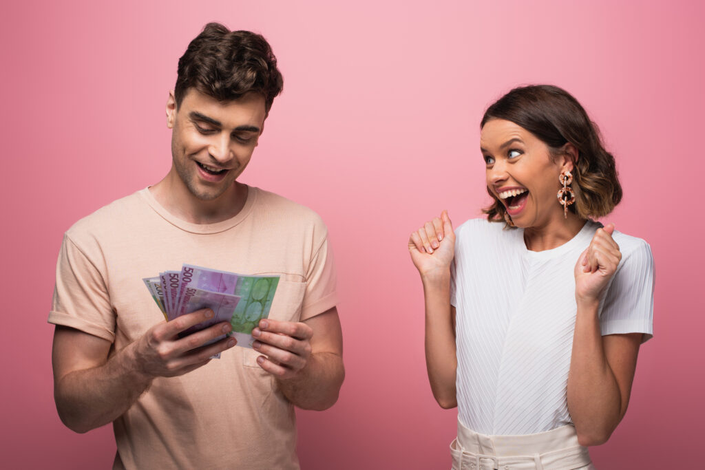 happy man counting dollar banknotes near excited girlfriend on pink background
