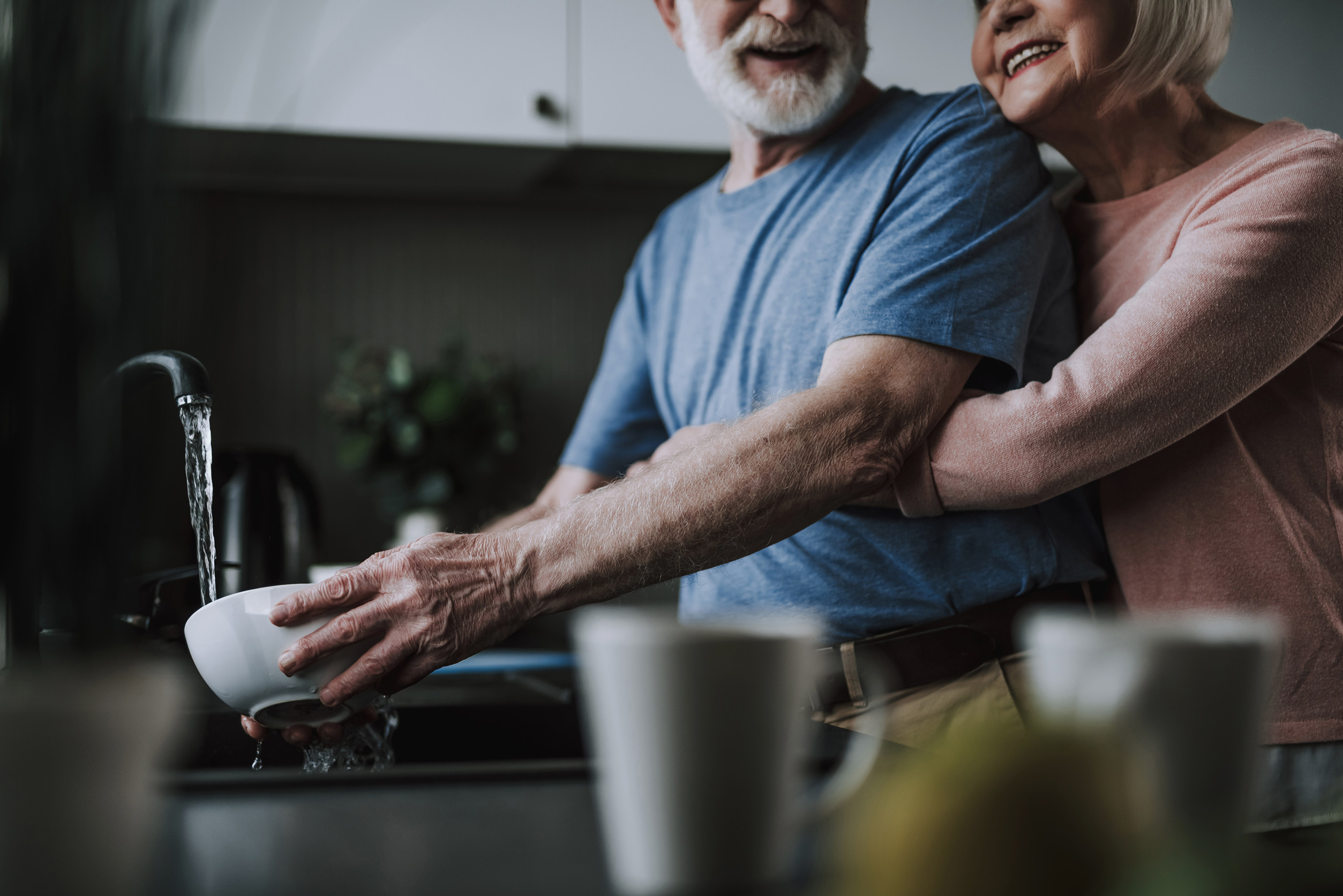 Enjoying time every moment. Waist up cropped head portrait of happy smiling senior man washing dishes while his wife tenderly embracing him from back