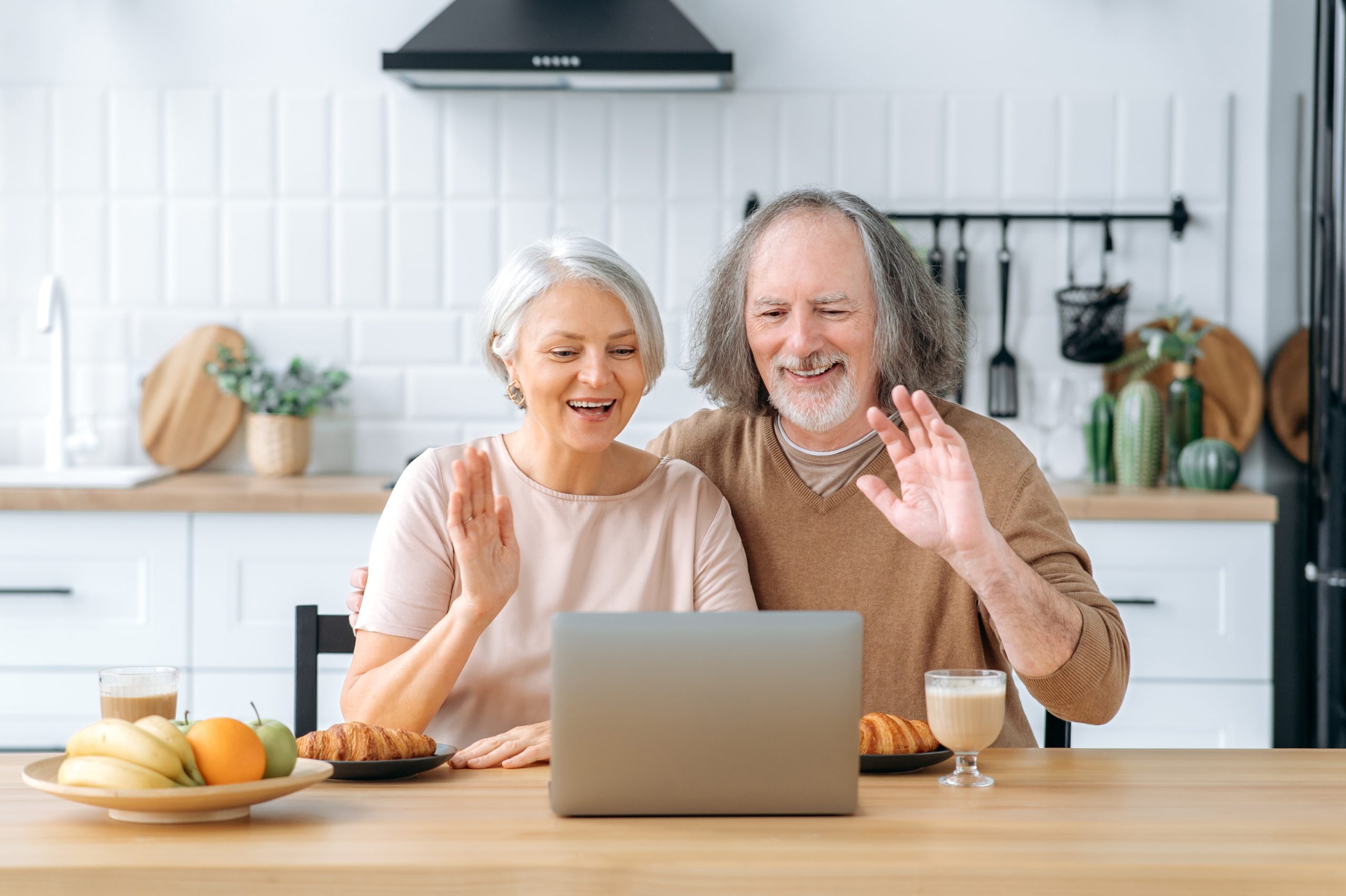 Happy caucasian pensioner couple, husband and wife, sit at home in the kitchen, use a laptop to communicate via online video conference with children and grandchildren, greet, smile joyfully