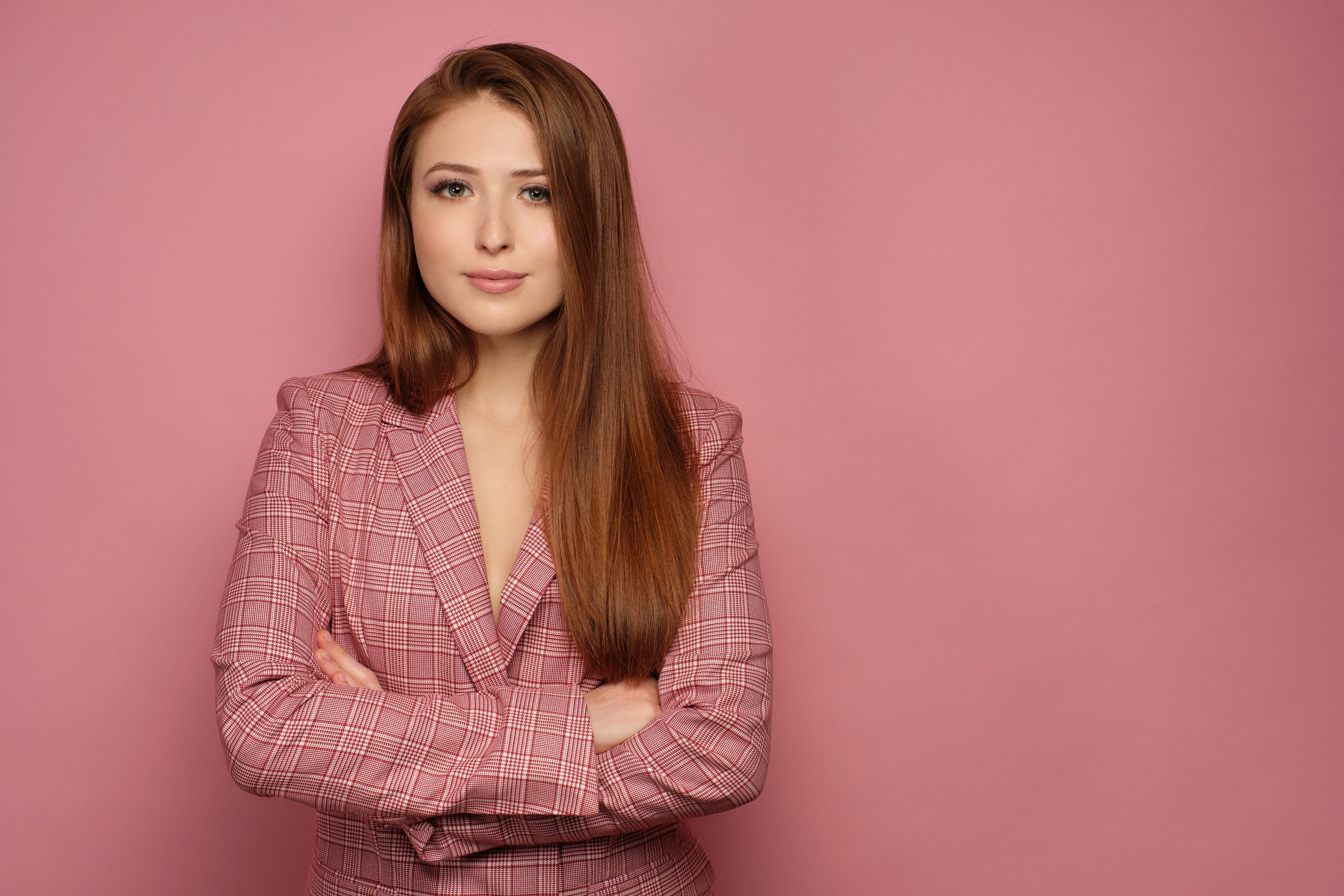 Red-haired girl in a pink business suit stands on a pink background with her arms folded and smiling looking at the camera. Free side space