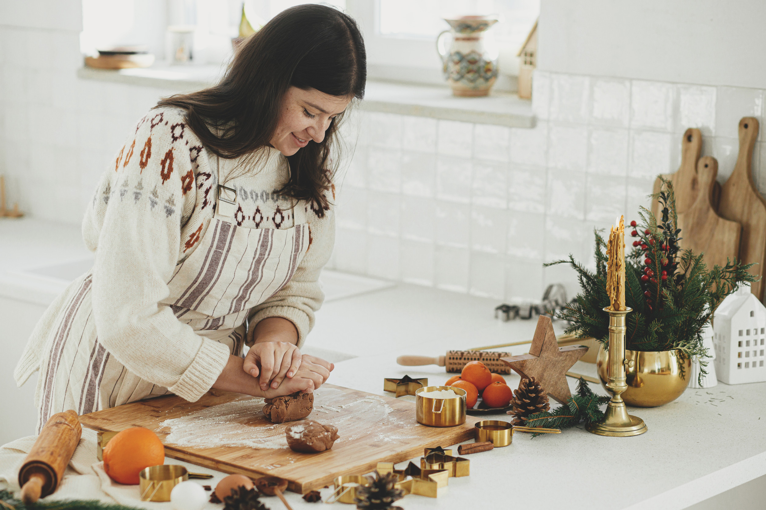 Woman making christmas gingerbread cookies in modern white kitchen. Hand kneading gingerbread dough on wooden board with flour, rolling pin, golden metal cutters, cooking spices on countertop
