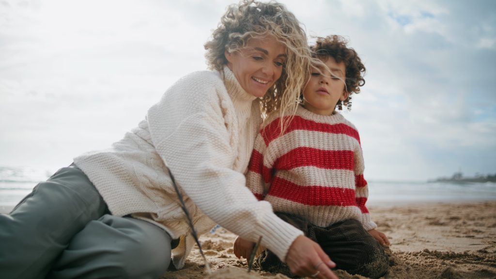 Curly mom kid building sand figures on ocean beach. Happy family relaxing weekend resting on cool autumn weekend together. Beautiful smiling mother babysitter play teaching kid. Shore holidays concept