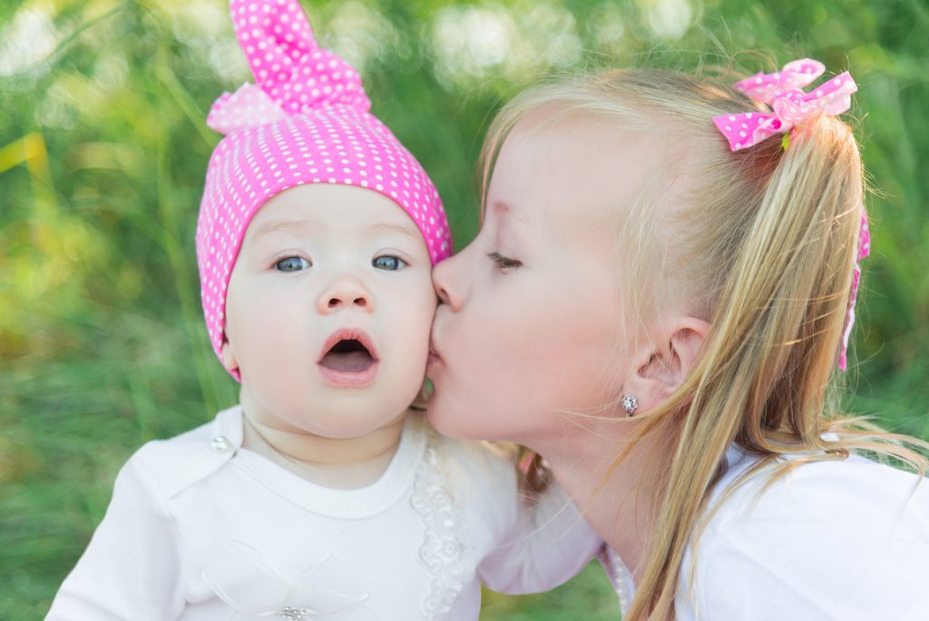 Two sisters. The older sister kisses the younger sister on the cheek. Pink hat in a pea and bows. Children are sitting in the grass. Children in the field.