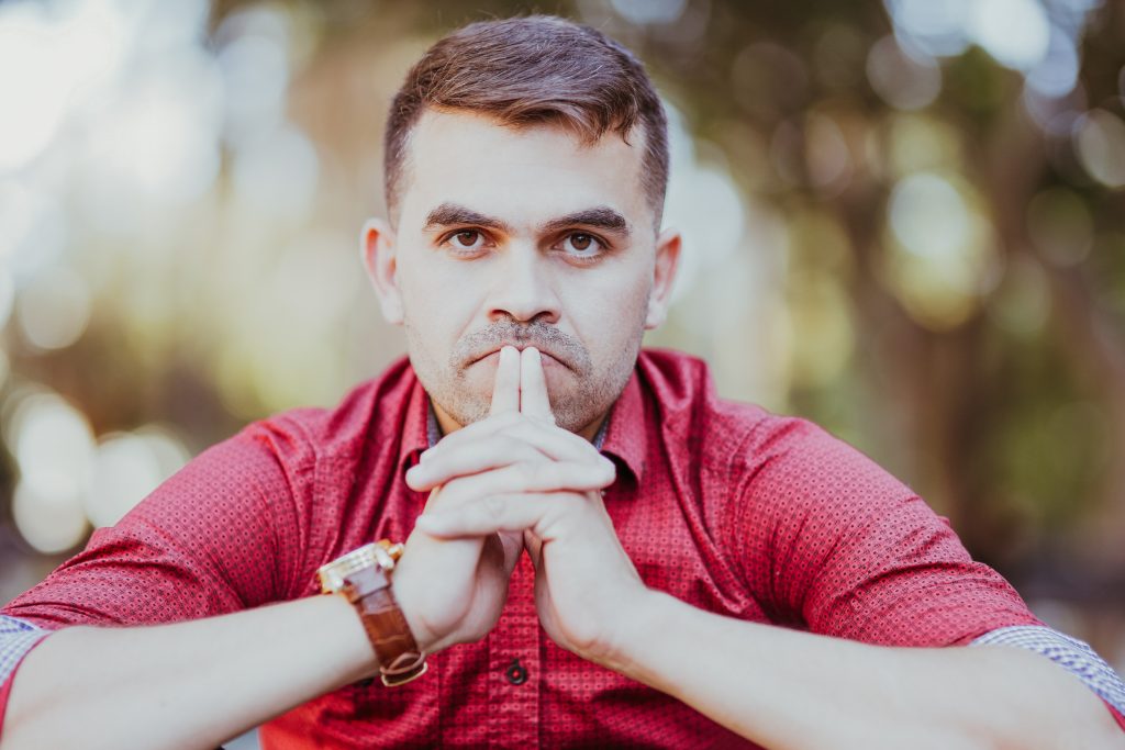 Portrait of pensive young Caucasian businessman wearing red shirt sitting, touching lips and looking at camera outdoors. Crisis and trouble concept