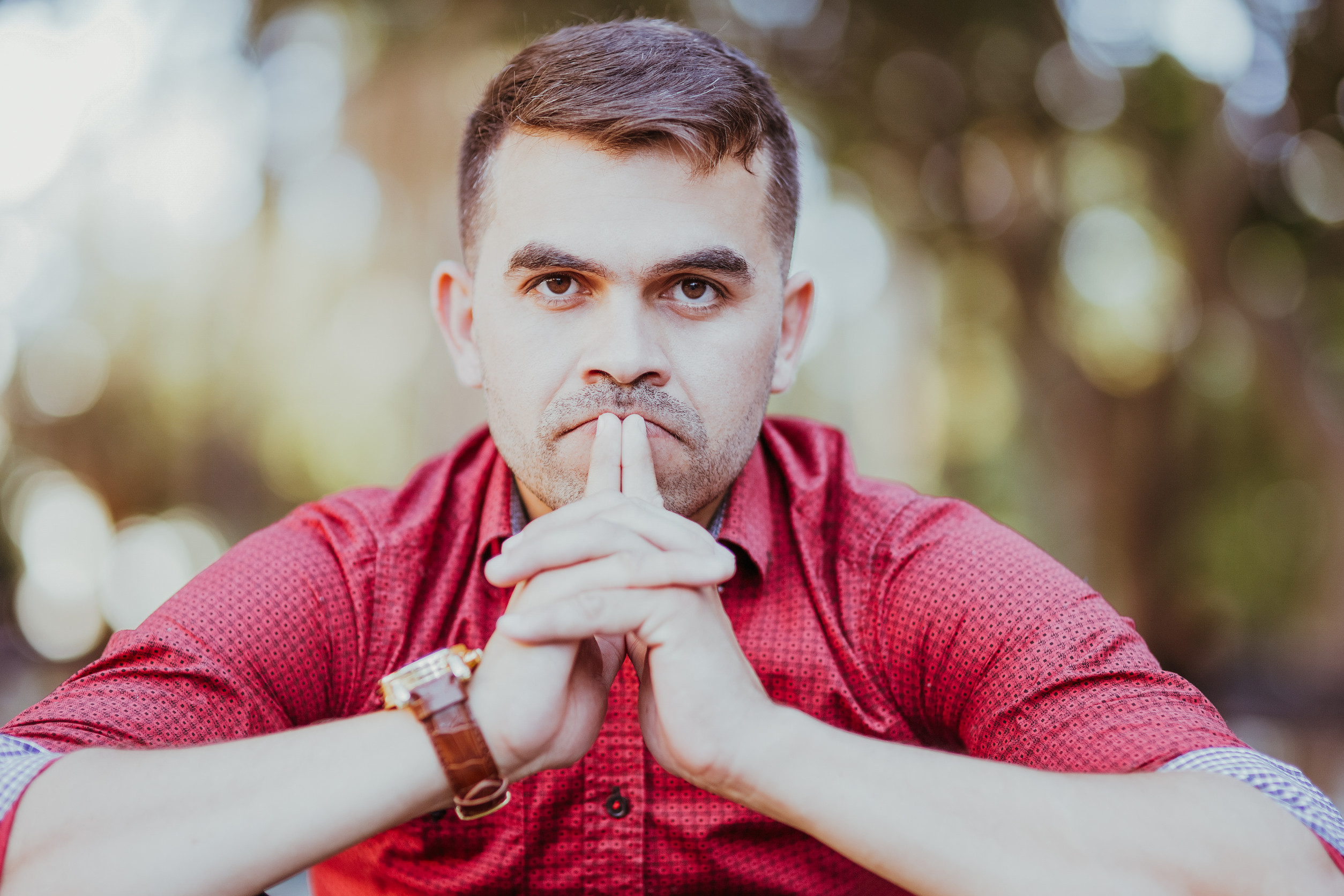 Portrait of pensive young Caucasian businessman wearing red shirt sitting, touching lips and looking at camera outdoors. Crisis and trouble concept
