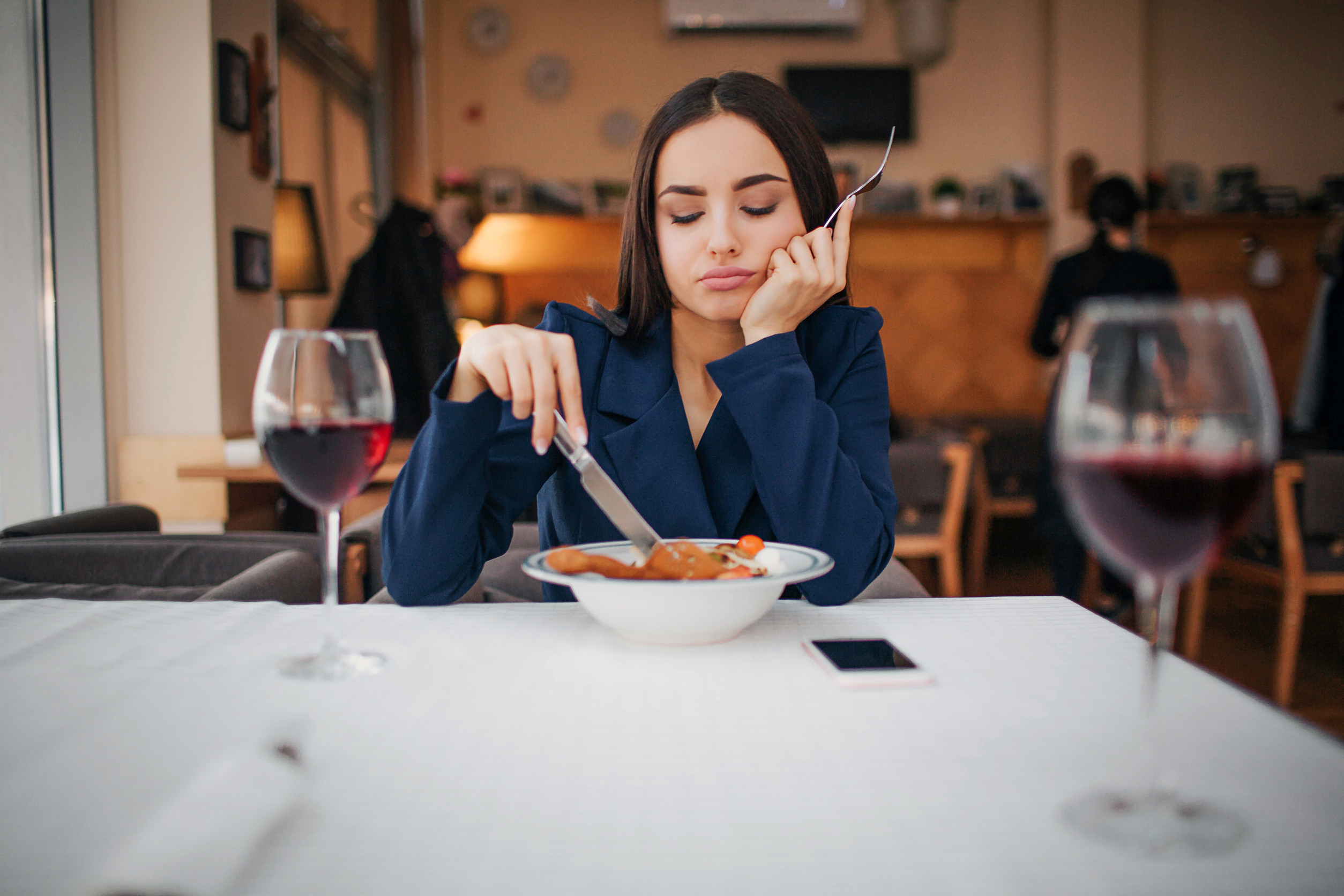 Bored young woman sit at table in restaurant alone. She look at food and touch it with fork. Two glasses of red wine stand on table