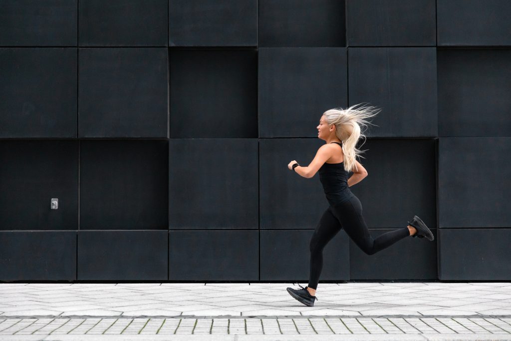 Side view of sporty young woman in black workout outfit running fast in minimalist urban environment. Sports woman with sport watch device.