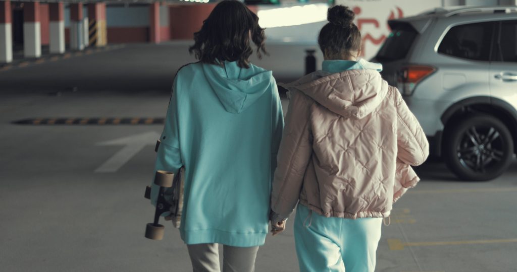 Two happy teen girls reunited in car parking hugging and walking home hand in hand. Urban city lifestyle. 4K video shooting by handheld gimbal