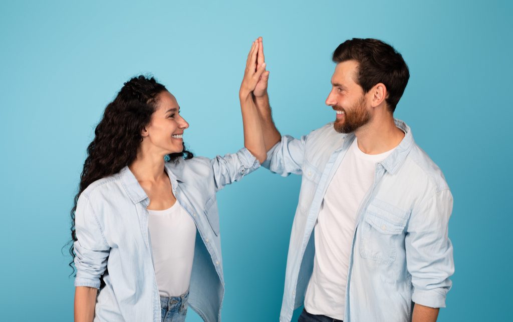 Smiling young european man and arab lady make high five hands sign, celebrating victory and success, have fun, isolated on blue background, studio, copy space. Emotions from win, good ad and offer