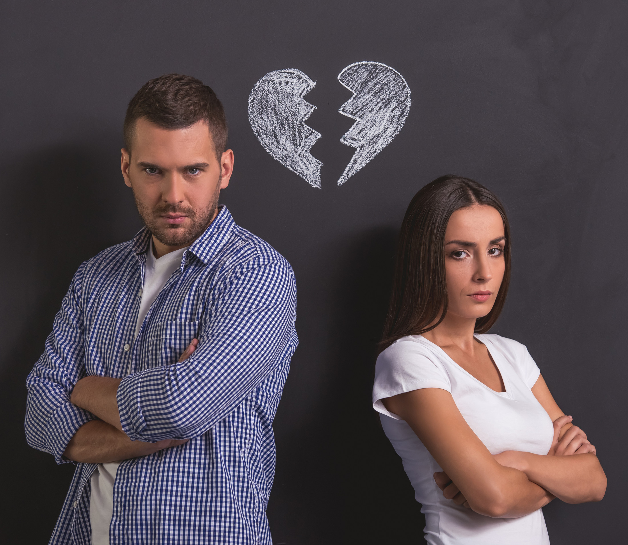 Beautiful young couple is looking at camera while standing back to back with folded arms against blackboard with drawn broken heart overhead